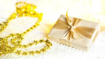 Box with Christmas gift in light paper is tied with a gold ribbon on white knitted background with decor. Golden garland of stars. New Year. Congratulations, surprise. space for text photo