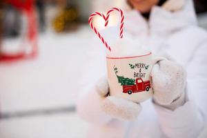 Woman with mug with snow, candy cane and inscription Merry and Bright in her hands outdoor in warm clothes in winter festive market. Fairy lights garlands decorated snow town for new year. Christmas photo