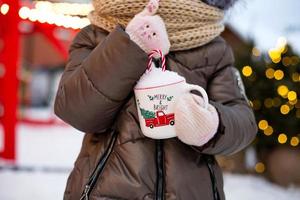 Girl with mug with snow, candy cane and inscription Merry and Bright in her hands outdoor in warm clothes in winter at festive market. Fairy lights garlands decorated snow town for new year. Christmas photo
