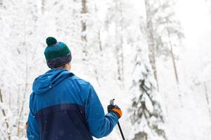 Skier in windbreaker and hat with pompom with ski poles in his hands with his back against the background of a snowy forest. Cross-country skiing in winter forest, outdoor sports, healthy lifestyle. photo