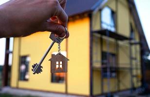 A hand with the keys to a new house on the background of an unfinished cottage. Building, project, moving to a new home, mortgage, rent and purchase real estate. To open the door. Copy space photo