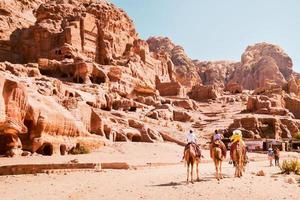 Local Arab tourist guide in Petra city riding on camel with tourists following group by royal Petra tombs in sunny hot day photo