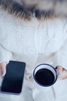 Top view and close up woman hands holding hot cup of coffee and smartphone in cold weather background, wearing warm fur knitted clothes photo