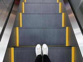 Selfie of feet in white sneakers shoes standing on escalator in shopping mall or modern office photo