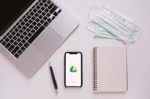 plano del escritorio del espacio de trabajo con teléfono inteligente que muestra la aplicación de Google Drive con máscara y texto covid19. google drive es un servicio que permite almacenar archivos en línea foto