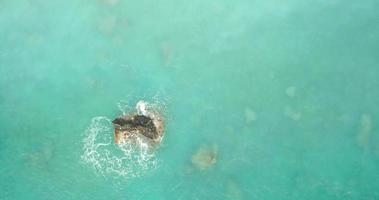 video de drones de una hermosa agua de mar turquesa y azul cristalina, rocas circundantes y olas que llegan a la playa de arena.