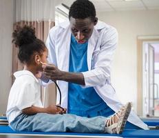 Selective focus at doctor person playing with cute afro child patient in hospital ward. Friendly pediatrician entertaining girl kid enjoying with stethoscope for heartbeat during medical exam. photo