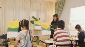 A female Asian teacher teaches and demonstrates to the children on acrylic color picture painting on canvas in art classroom, creatively learning with skill at the elementary school studio education. video