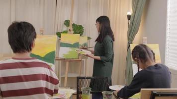 A female Asian teacher teaches and demonstrates to the children on acrylic color picture painting on canvas in art classroom, creatively learning with skill at the elementary school studio education. video