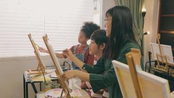 A female Asian teacher teaches and demonstrates to the children on acrylic color picture painting on canvas in art classroom, creatively learning with skill at the elementary school studio education. video