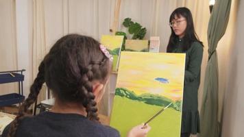 Female Asian teacher teaches and demonstrates to little student girl on acrylic color picture painting on canvas in art classroom, creatively learning with skill at elementary school studio education. video