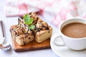 Delicioso postre de pastel de café servido en la mesa - rebanada de pastel de chocolate sobre fondo de tablero de madera con hoja de menta y taza de café para el desayuno foto