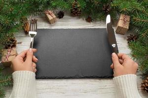 vista en perspectiva la niña sostiene un tenedor y un cuchillo en la mano y está lista para comer. placa rectangular de pizarra negra vacía sobre fondo de Navidad de madera. concepto de plato de cena navideña con decoración de año nuevo foto