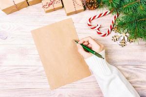 chica mano escribiendo carta de navidad en papel artesanal con decoraciones sobre fondo de madera. tonificado foto