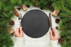 top view girl holds fork and knife in hand and is ready to eat. Empty black slate plate on wooden christmas background. holiday dinner dish concept with new year decor photo