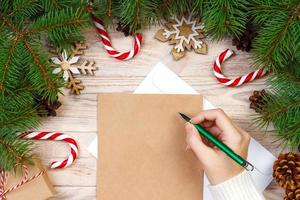 chica mano escribiendo carta de navidad en papel artesanal con decoraciones sobre fondo de madera foto