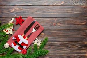 Top view of fork and knife put in santa clothes on napkin with christmas decorations and new year tree on wooden background. Holiday and festive concept with copy space photo