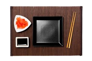 Empty black square plate with chopsticks for sushi, ginger and soy sauce on dark bamboo mat background. Top view with copy space for you design photo