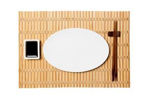 plato blanco ovalado vacío con palillos para sushi y salsa de soja sobre fondo de estera de bambú amarillo. vista superior con espacio de copia para su diseño foto