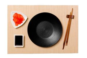 Empty round black plate with chopsticks for sushi, ginger and soy sauce on brown sushi mat background. Top view with copy space for you design photo