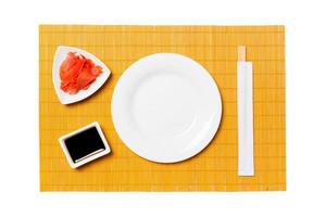 Empty round white plate with chopsticks for sushi and soy sauce, ginger on yellow bamboo mat background. Top view with copy space for you design photo