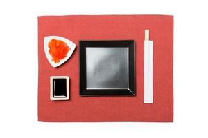 Empty black square plate with chopsticks for sushi, ginger and soy sauce on red napkin background. Top view with copy space for you design photo