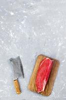 fresh large piece of beef on a wooden cutting board with kitchen ax. Top view on gray concrete background photo