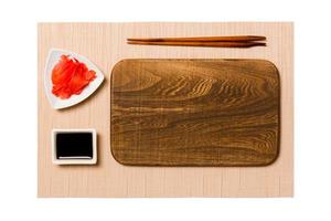 Empty rectangular brown wooden plate with chopsticks for sushi, ginger and soy sauce on brown sushi mat background. Top view with copy space for you design photo