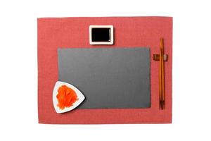 Empty rectangular black slate plate with chopsticks for sushi, ginger and soy sauce on red napkin background. Top view with copy space for you design photo