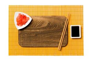 Empty rectangular brown wooden plate with chopsticks for sushi, ginger and soy sauce on yellow bamboo mat background. Top view with copy space for you design photo