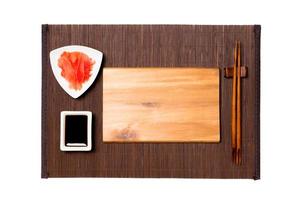 Empty rectangular brown wooden plate with chopsticks for sushi, ginger and soy sauce on dark bamboo mat background. Top view with copy space for you design photo