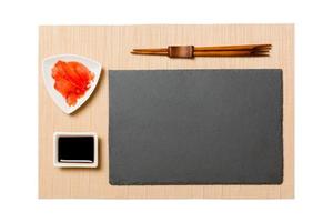 Empty rectangular black slate plate with chopsticks for sushi, ginger and soy sauce on brown sushi mat background. Top view with copy space for you design photo