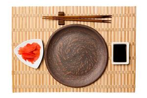 Empty round brown plate with chopsticks for sushi and soy sauce, ginger on yellow bamboo mat background. Top view with copy space for you design photo