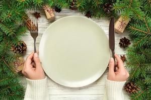 top view girl holds fork and knife in hand and is ready to eat. Empty plate round ceramic on wooden christmas background. holiday dinner dish concept with new year decor photo