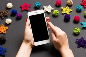 Top view of phone in female hand on festive black background. Christmas decorations. New Year time holiday. Mockup photo