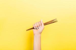 Cropped image of female hand holding chopsticks in fist on yellow background. Asian food concept with copy space photo