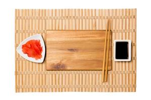 Empty rectangular brown wooden plate with chopsticks for sushi and soy sauce, ginger on yellow bamboo mat background. Top view with copy space for you design photo