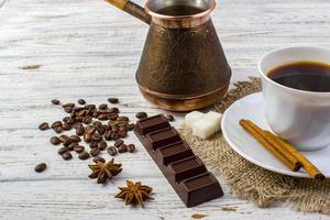 A white cup with black coffee on sacking, anise, sugar, a piece of chocolate, coffee beans and cinnamon sticks on bright wooden table photo