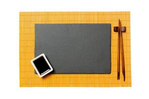 placa de pizarra negra rectangular vacía con palillos para sushi y salsa de soja sobre fondo de estera de bambú amarillo. vista superior con espacio de copia para su diseño foto