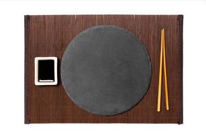 Emptyround black slate plate with chopsticks for sushi and soy sauce on dark bamboo mat background. Top view with copy space for you design photo