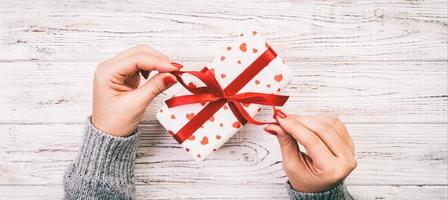 Woman hands give wrapped valentine or other holiday handmade present in paper with red ribbon. Present box, red heart of gift on table, Top view banner with copy space for you design. Toned web banner photo