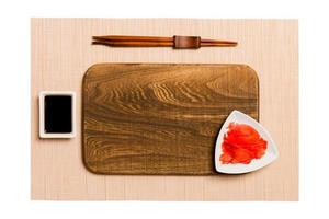 Empty rectangular brown wooden plate with chopsticks for sushi, ginger and soy sauce on brown sushi mat background. Top view with copy space for you design photo