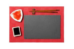 Empty rectangular black slate plate with chopsticks for sushi, ginger and soy sauce on red mat sushi background. Top view with copy space for you design photo