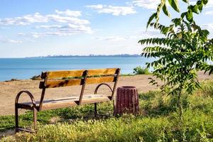 A simple minimalistic layout of a bench on green grass looking out toward the ocean sea beach waters with blue blank empty copyspace sky above the horizon photo