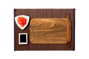 Empty rectangular brown wooden plate with chopsticks for sushi, ginger and soy sauce on dark bamboo mat background. Top view with copy space for you design photo