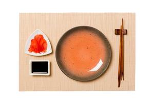 Empty round brown plate with chopsticks for sushi, ginger and soy sauce on brown sushi mat background. Top view with copy space for you design photo