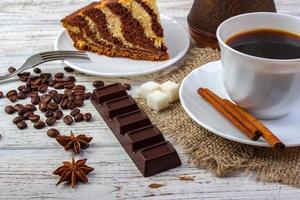 A white cup with coffee on sacking and a piece of amazing chocolate cake on a plate with a fork. A chocolate bar, coffee beans, anice, cinnamon sticks, sugar on wooden background photo