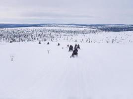 A group of tourists riding Snowmobiles on Glacier through the snowy mountains in Finland, panoramic scene of white snow hills with Pine Tree. photo