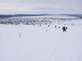 A group of tourists riding Snowmobiles on Glacier through the snowy mountains in Finland, panoramic scene of white snow hills with Pine Tree. photo