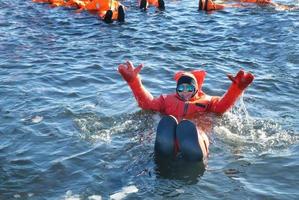 A woman in sunglasses raised both arms swimming in cold ice water pond with orange color of water right thermal suit, tourist adventure activity, Arctic Circle photo